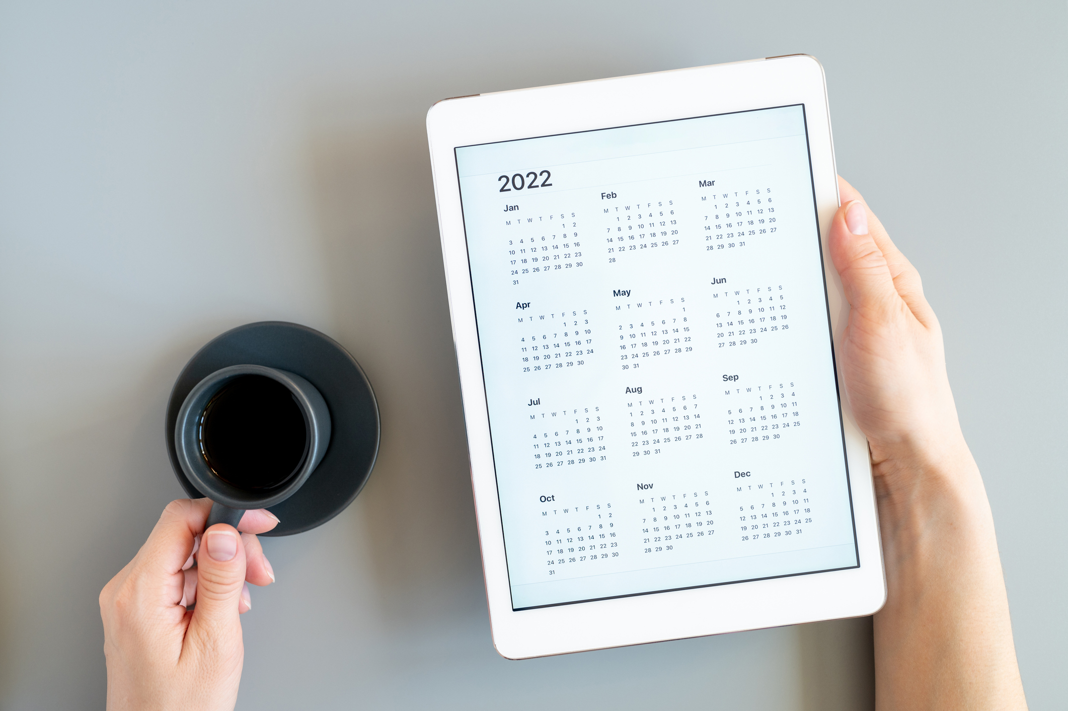 Person holding a tablet displaying a yearly calendar while drinking coffee, symbolizing year-end procurement planning and process streamlining.