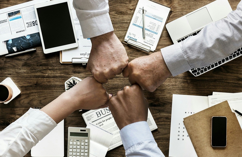 Team members joining fists over a desk filled with business documents and devices, symbolizing collaborative support for implementing digital procurement.