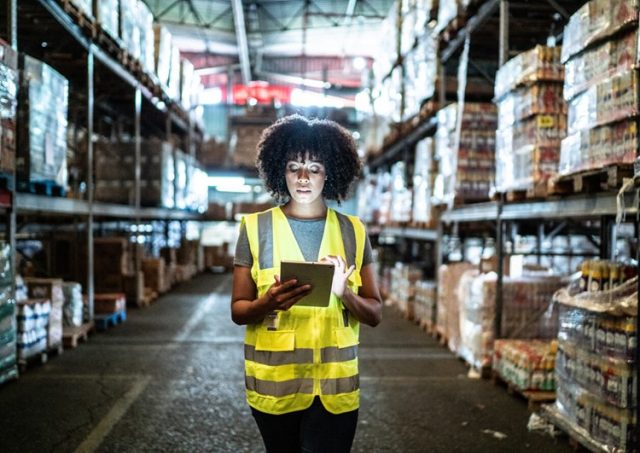 Warehouse worker wearing a safety vest using a tablet while standing between stocked shelves.